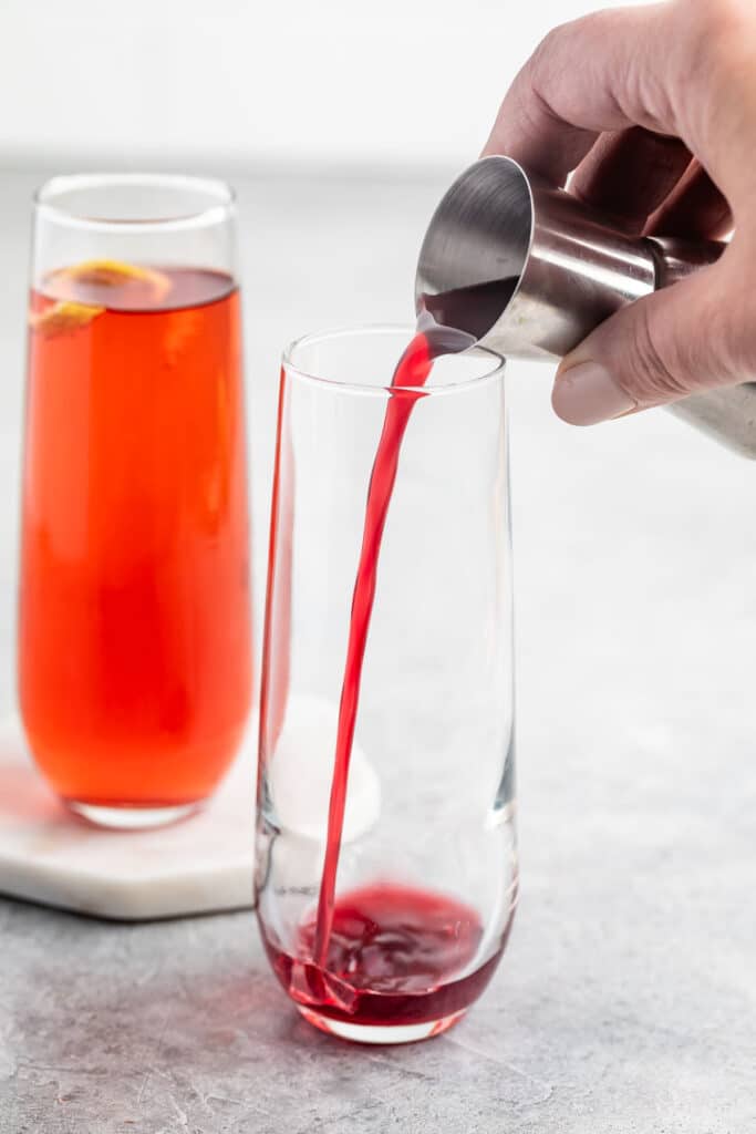 A person pours a bright red liquid from a measuring cup into a clear glass. Another glass with a similar red drink and an orange slice is in the background on a coaster. The surface is a light gray.