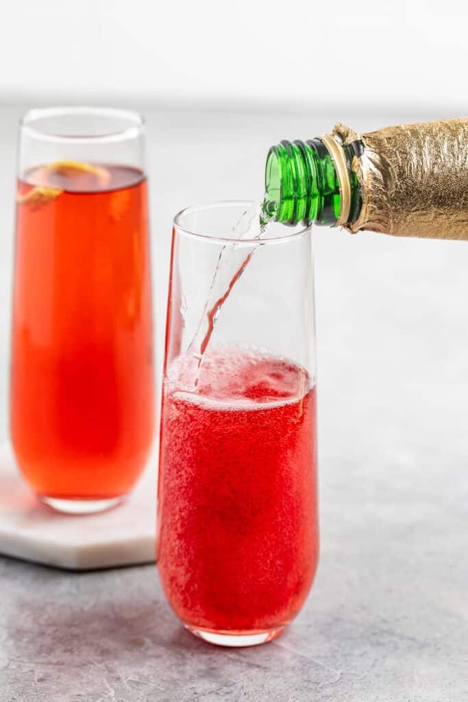 A close-up of two tall glasses filled with a bright red beverage. One glass is being filled with a drink from a green bottle wrapped in gold foil. A slice of lemon is visible in the other glass, which rests on a coaster.