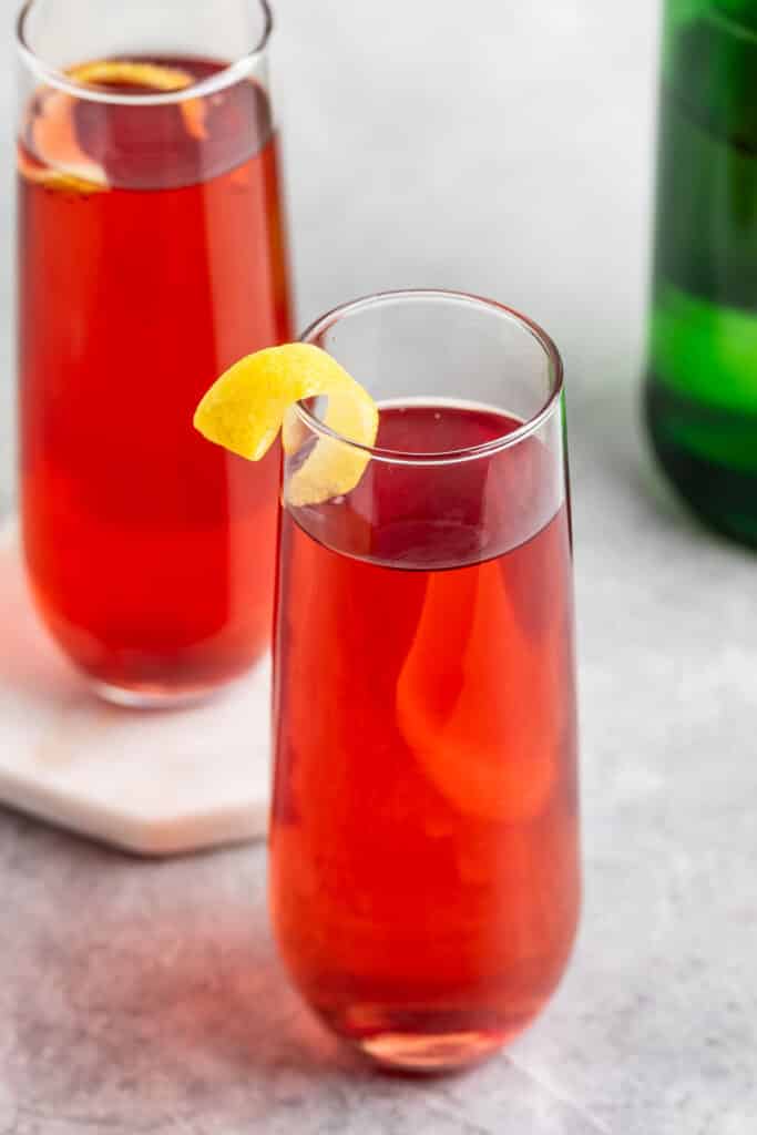 Two tall glasses filled with a red cocktail are garnished with lemon twists, set on a light gray surface. In the background, a green bottle is partially visible.