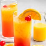 Two tall glasses of colorful cocktails with orange slices and cherries garnish the rim, surrounded by scattered orange slices and cherries. A small bottle of orange juice is in the background on a light gray surface.