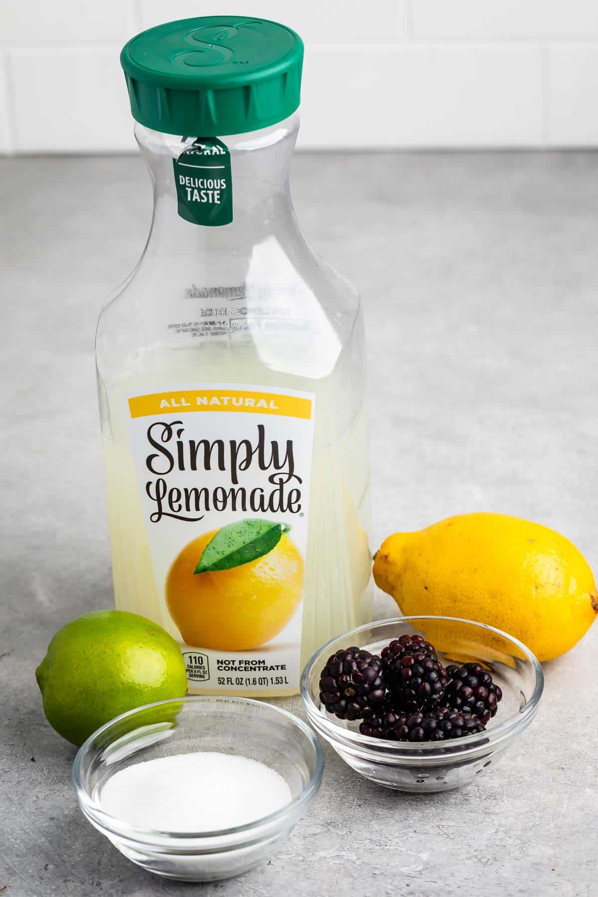 A bottle of Simply Lemonade is surrounded by a lemon, a lime, a bowl of blackberries, and a bowl of sugar. All items are placed on a gray surface with a white tiled background.