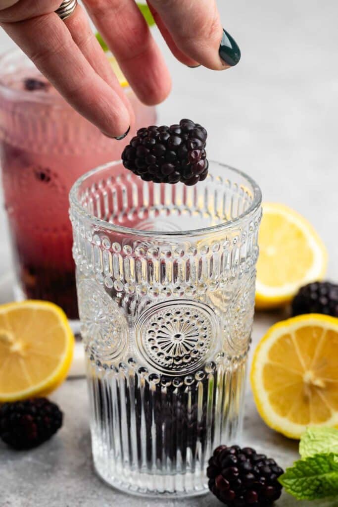 A hand drops a blackberry into an ornate glass of berry-infused water; another glass and sliced lemons are in the background, with fresh blackberries and mint leaves scattered on a gray surface.