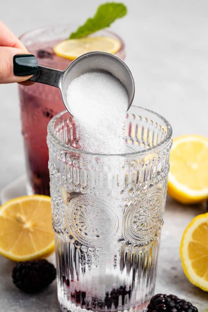 A hand pours sugar into a textured glass filled with a purple beverage. Slices of lemon and blackberries surround the glass on a gray surface. Another drink with a mint leaf is in the background.