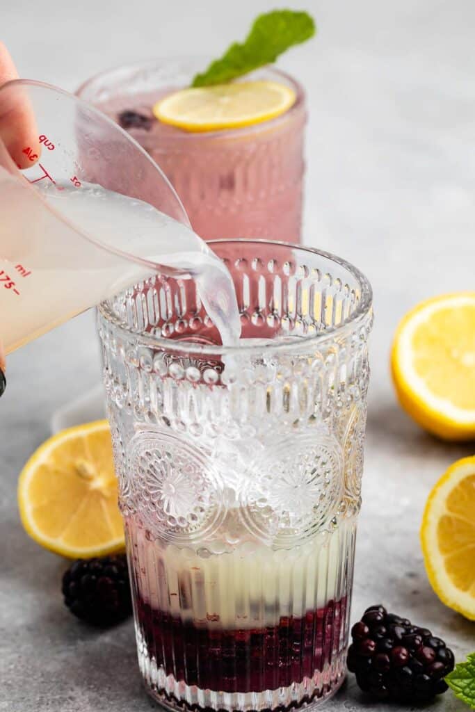 A hand pours a clear liquid into a patterned glass containing a dark berry mixture and ice. Another similar glass, garnished with a lemon slice and mint, is in the background. Lemon halves and blackberries surround the glasses on the table.