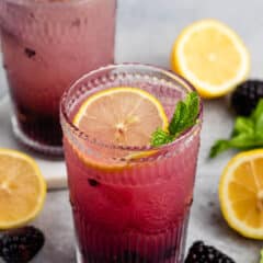 Two glasses of refreshing blackberry lemonade garnished with lemon slices and mint leaves. Surrounding the glasses are fresh blackberries, lemon halves, and mint sprigs, all set on a light gray surface.