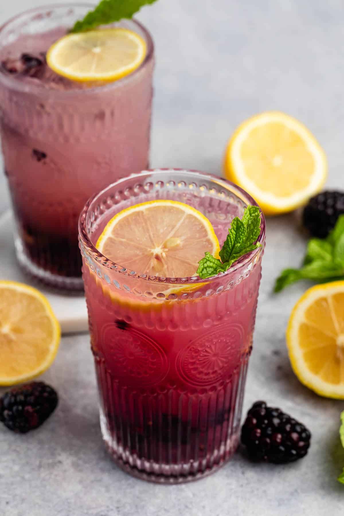 Two glasses of refreshing blackberry lemonade garnished with lemon slices and mint leaves. Surrounding the glasses are fresh blackberries, lemon halves, and mint sprigs, all set on a light gray surface.