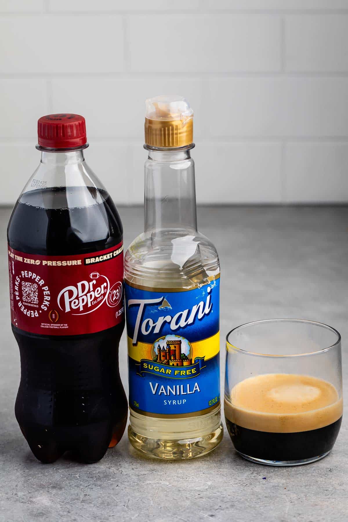 A bottle of Dr Pepper, a bottle of Torani Sugar Free Vanilla Syrup, and a glass with a dark beverage are arranged on a gray countertop with a white tile background.