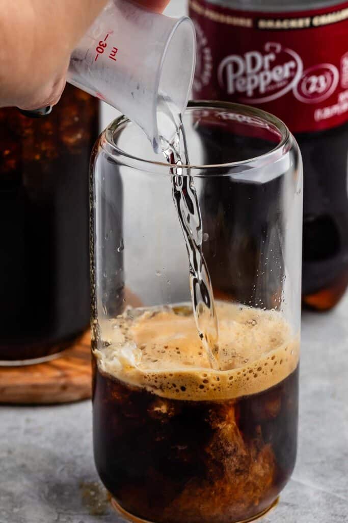 A clear glass is being filled with a fizzy liquid from a small measuring cup. A bottle of Dr. Pepper is visible in the background. The drink inside the glass is dark and foamy.