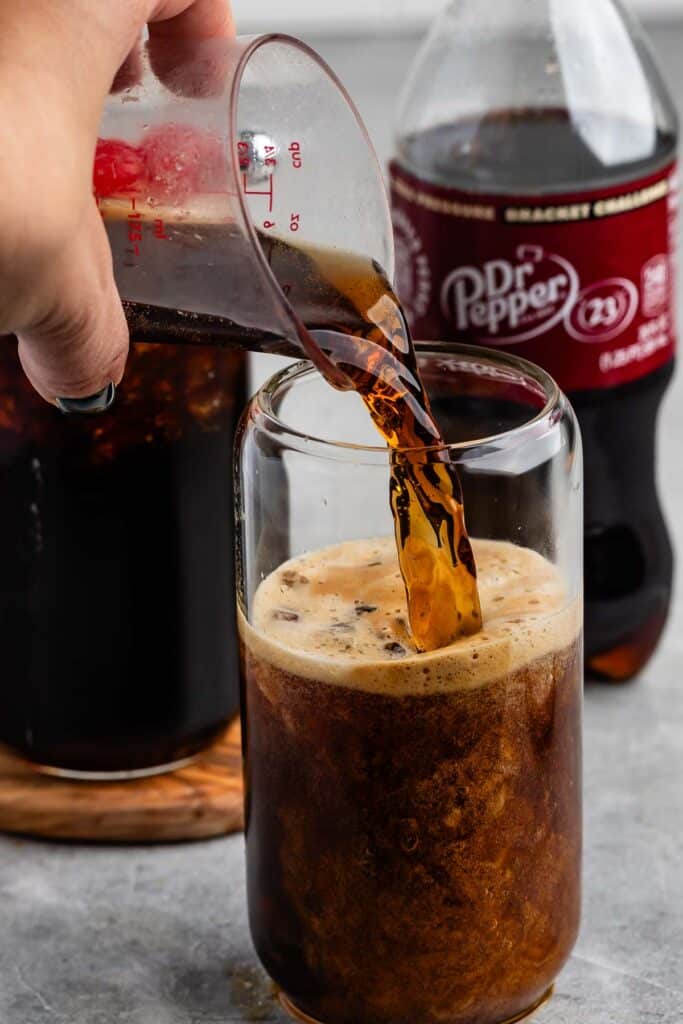 A hand pours Dr. Pepper from a measuring cup into a glass with ice. A bottle of Dr. Pepper stands in the background.