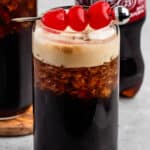 A glass of dark soda with a frothy top is garnished with three red cherries on a skewer. In the background, a bottle of Dr Pepper is partially visible. The glass sits on a gray surface.