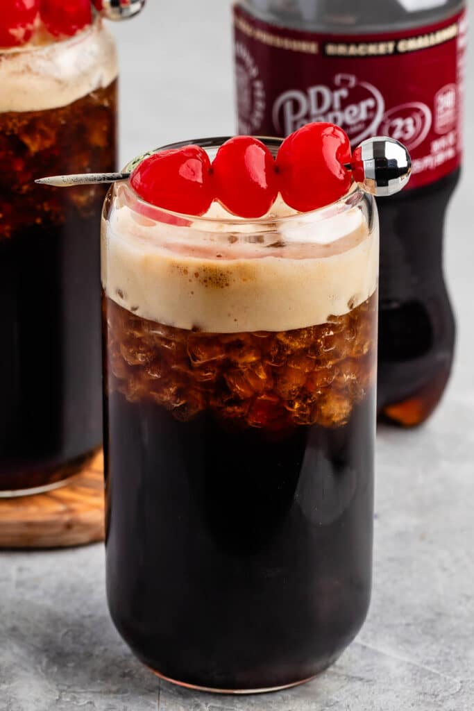 A glass of dark soda with a frothy top is garnished with three red cherries on a skewer. In the background, a bottle of Dr Pepper is partially visible. The glass sits on a gray surface.