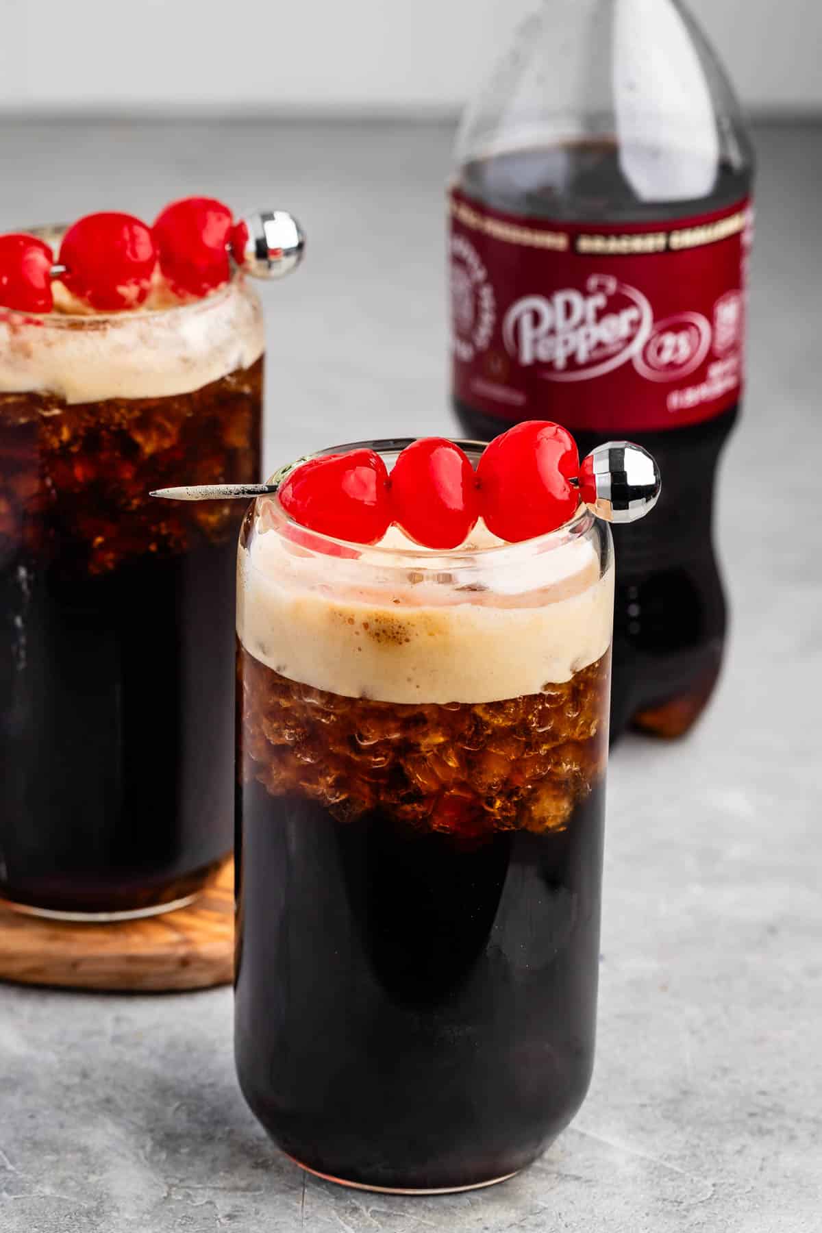 Two tall glasses filled with a dark soda and topped with foam are garnished with red cherries on skewers. A bottle of Dr Pepper is in the background on a gray surface.