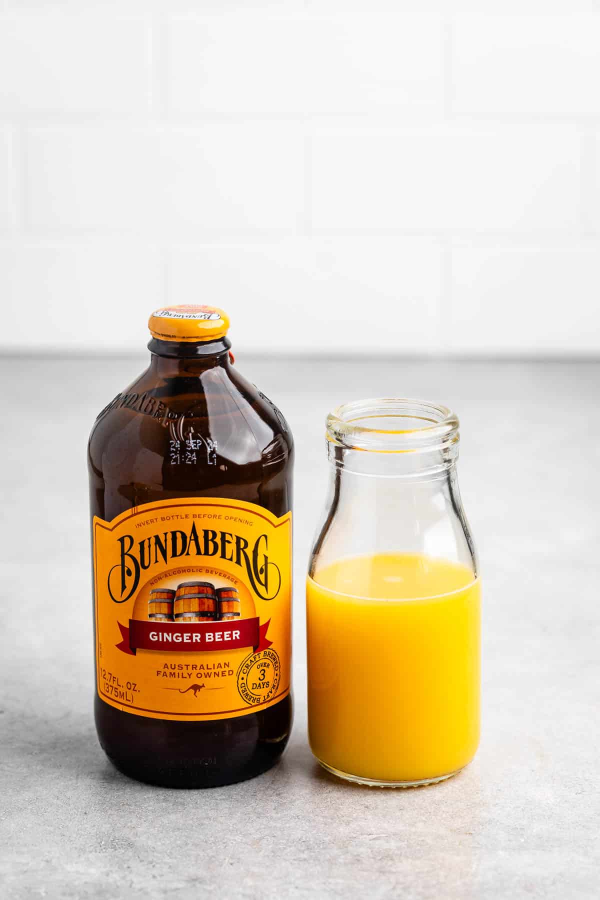 A bottle of Bundaberg ginger beer stands next to a glass bottle filled with orange liquid on a gray surface, set against a white tile background.