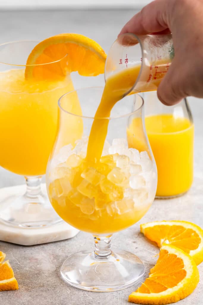 A hand pours orange juice over ice into a glass. Another glass with orange juice and a slice of orange on the rim sits in the background next to a small bottle of juice and slices of fresh orange on a gray countertop.