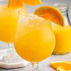 Two glasses of bright orange juice with crushed ice, garnished with orange slices, are placed on a light gray surface. A small glass bottle of juice is in the background, and fresh orange slices are scattered around.