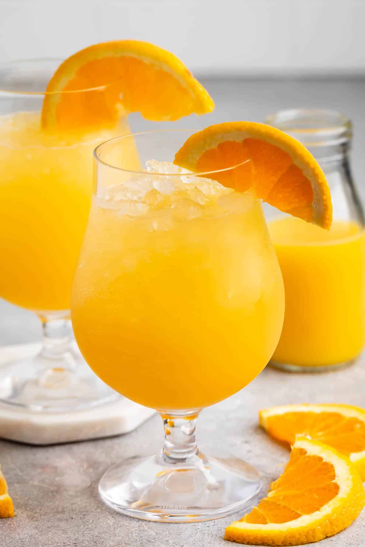 Two glasses of bright orange juice with crushed ice, garnished with orange slices, are placed on a light gray surface. A small glass bottle of juice is in the background, and fresh orange slices are scattered around.