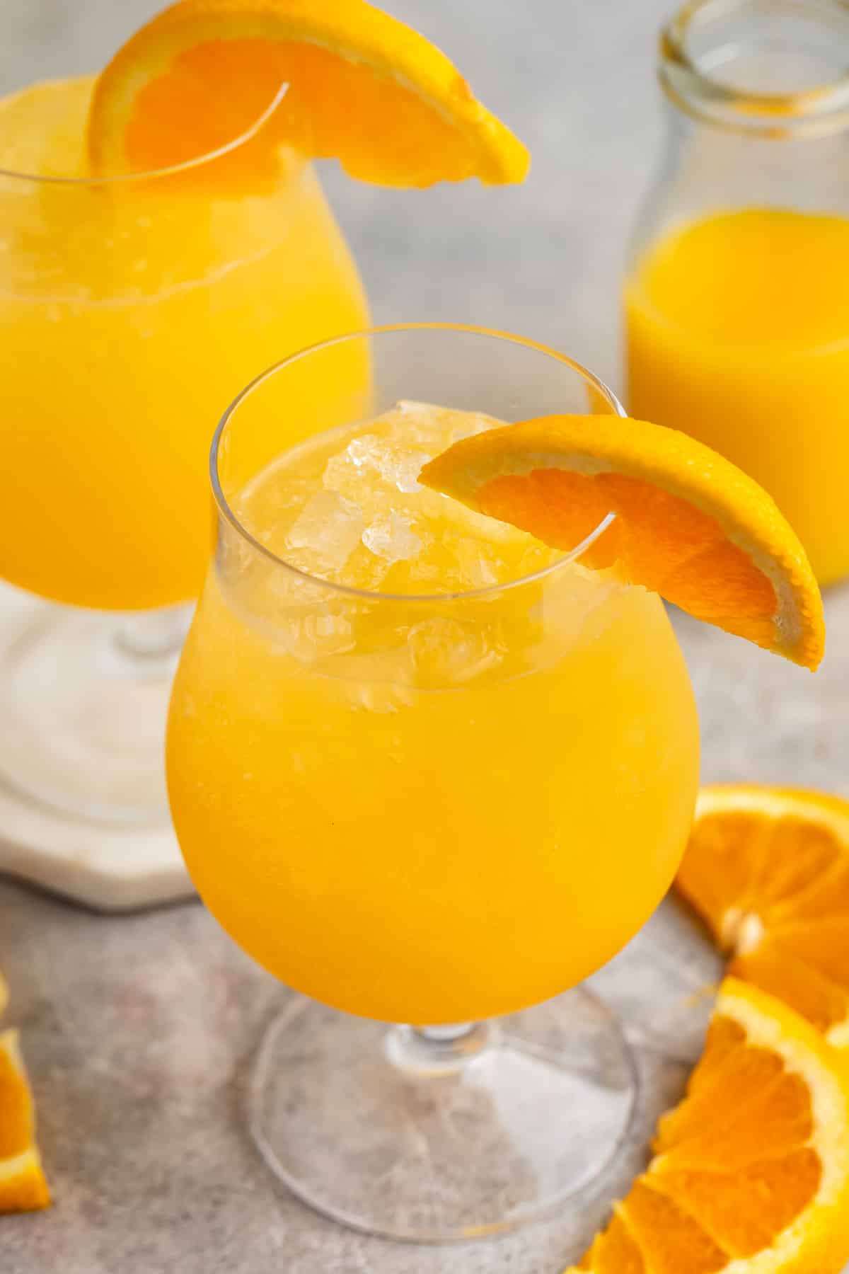 A glass of orange juice with ice and a slice of orange on the rim sits on a table. A second glass and a small bottle of juice are in the background. Orange slices are scattered on the table surface.