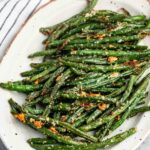 A white oval plate filled with roasted green beans sprinkled with grated cheese and herbs. The background features a striped fabric.