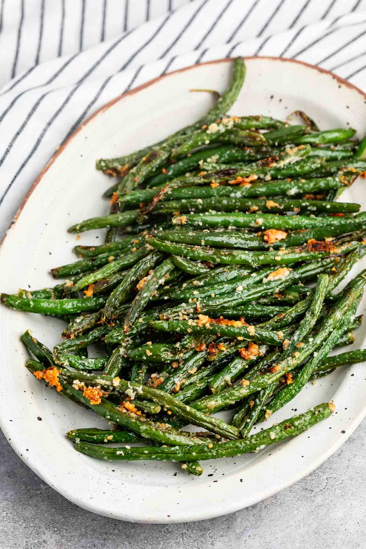 A white oval plate filled with roasted green beans sprinkled with grated cheese and herbs. The background features a striped fabric.