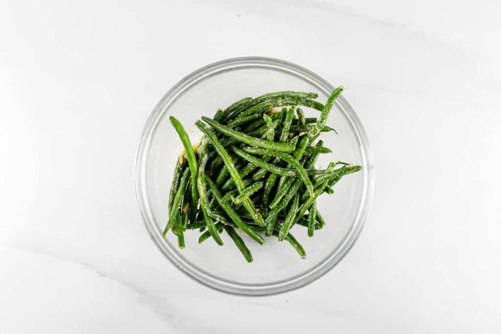 A glass bowl filled with cooked green beans on a white marble surface. The green beans are seasoned and glistening, suggesting they are freshly prepared. The overhead view highlights their vibrant green color and texture.