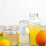 Three glass jars filled with different liquids are displayed on a surface. Surrounding them are lemons, a lime, an orange, and sprigs of rosemary. A white tiled wall serves as the background.