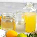 Three glass bottles containing different liquids are placed on a countertop. In front of them are lemons, a lime, a bowl of sugar, and sprigs of rosemary. The background is a white tiled wall.