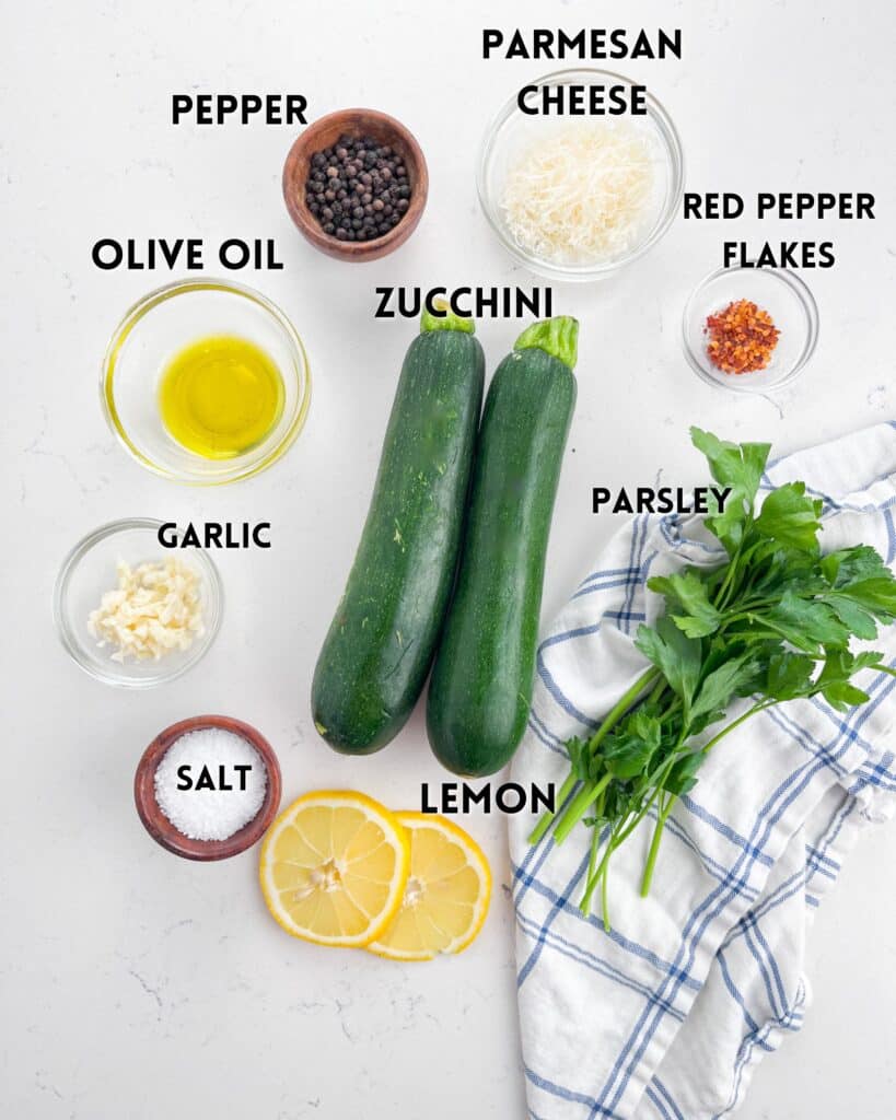 Ingredients for a recipe labeled on a white surface, including zucchini, garlic, salt, whole black pepper, olive oil, grated Parmesan cheese, lemon slices, red pepper flakes, and parsley, arranged around a blue-striped cloth.