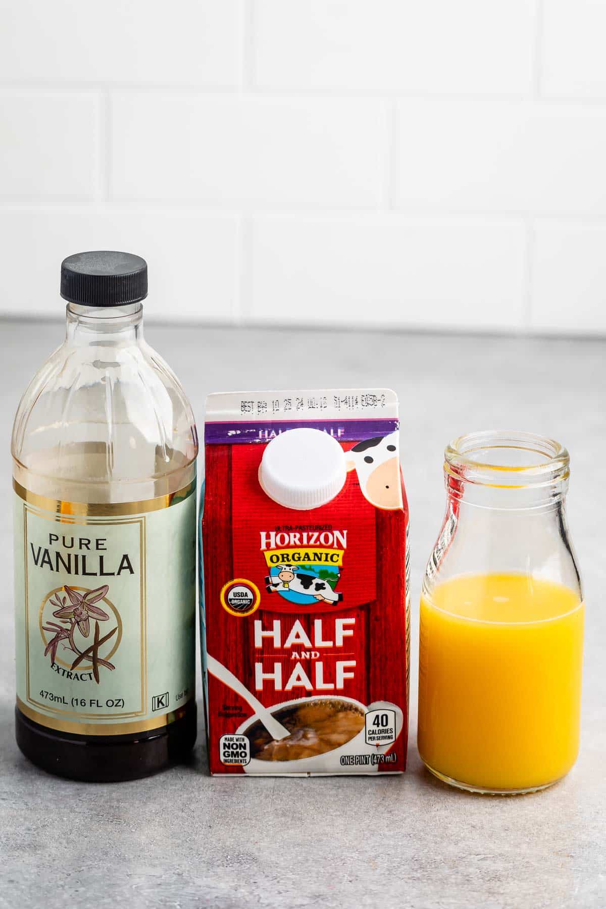 A bottle of pure vanilla extract, a carton of Horizon Organic half and half, and a glass bottle filled with orange juice are placed on a gray countertop against a white tile background.