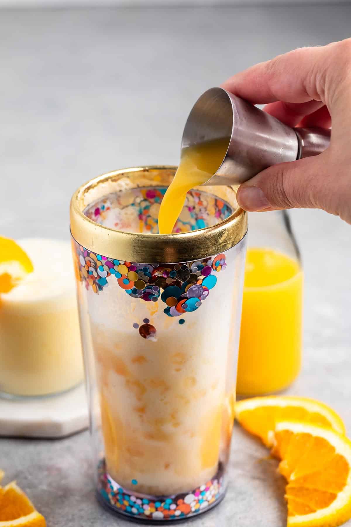 A hand pours orange juice from a small metal measuring cup into a transparent tumbler filled with ice. The tumbler has colorful decorations. Slices of orange and a glass of juice are visible in the background.