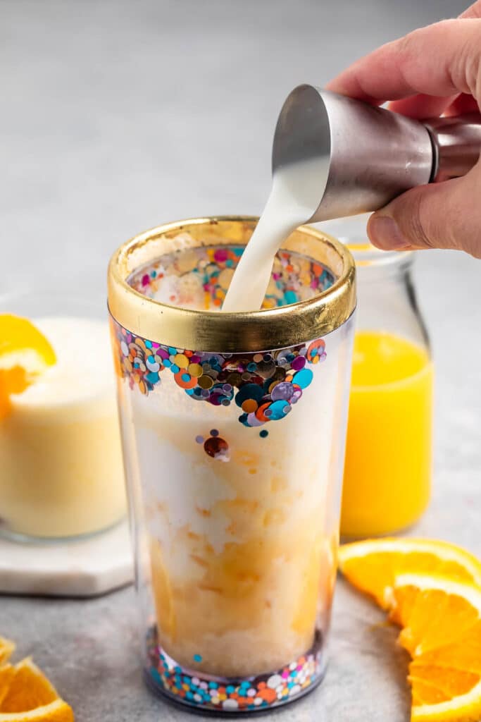 A hand pours milk from a small metal cup into a glitter-decorated tumbler filled with a creamy beverage. Nearby are fresh orange slices, a glass of orange juice, and an orange dessert on a white coaster.