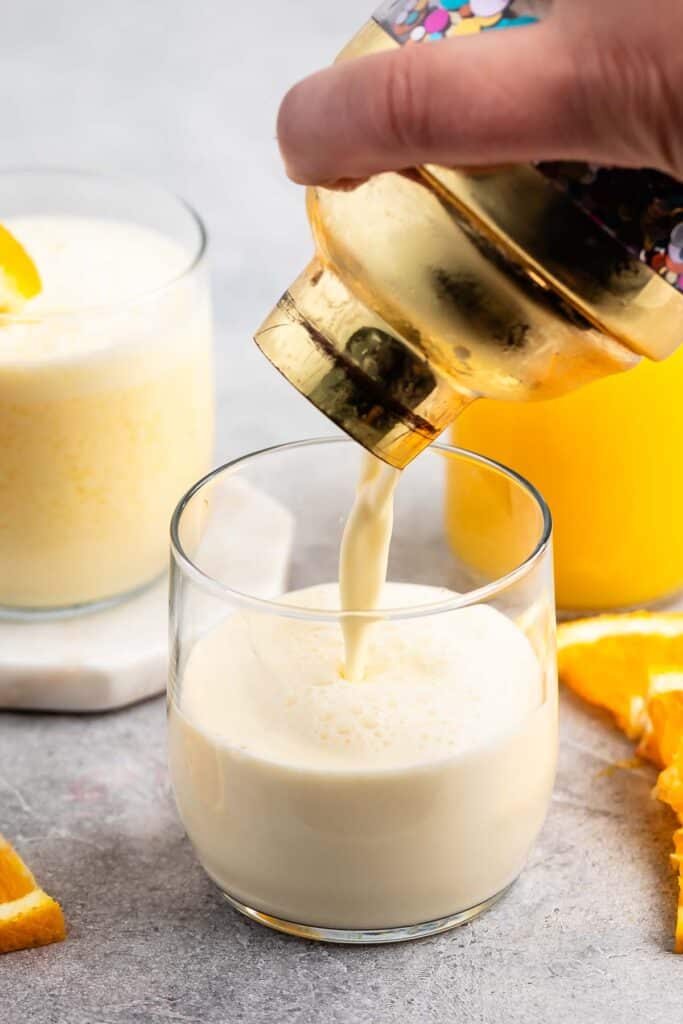 A hand pours a creamy, frothy drink from a shaker into a glass. In the background, there is another filled glass and a pitcher of orange juice. Orange slices are scattered on the gray surface.