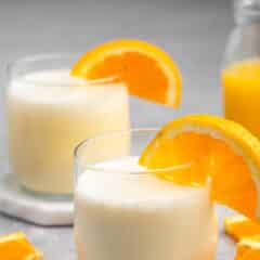 Two glasses of creamy orange smoothies garnished with orange slices. A bottle of orange juice and additional orange slices are in the background on a gray surface.