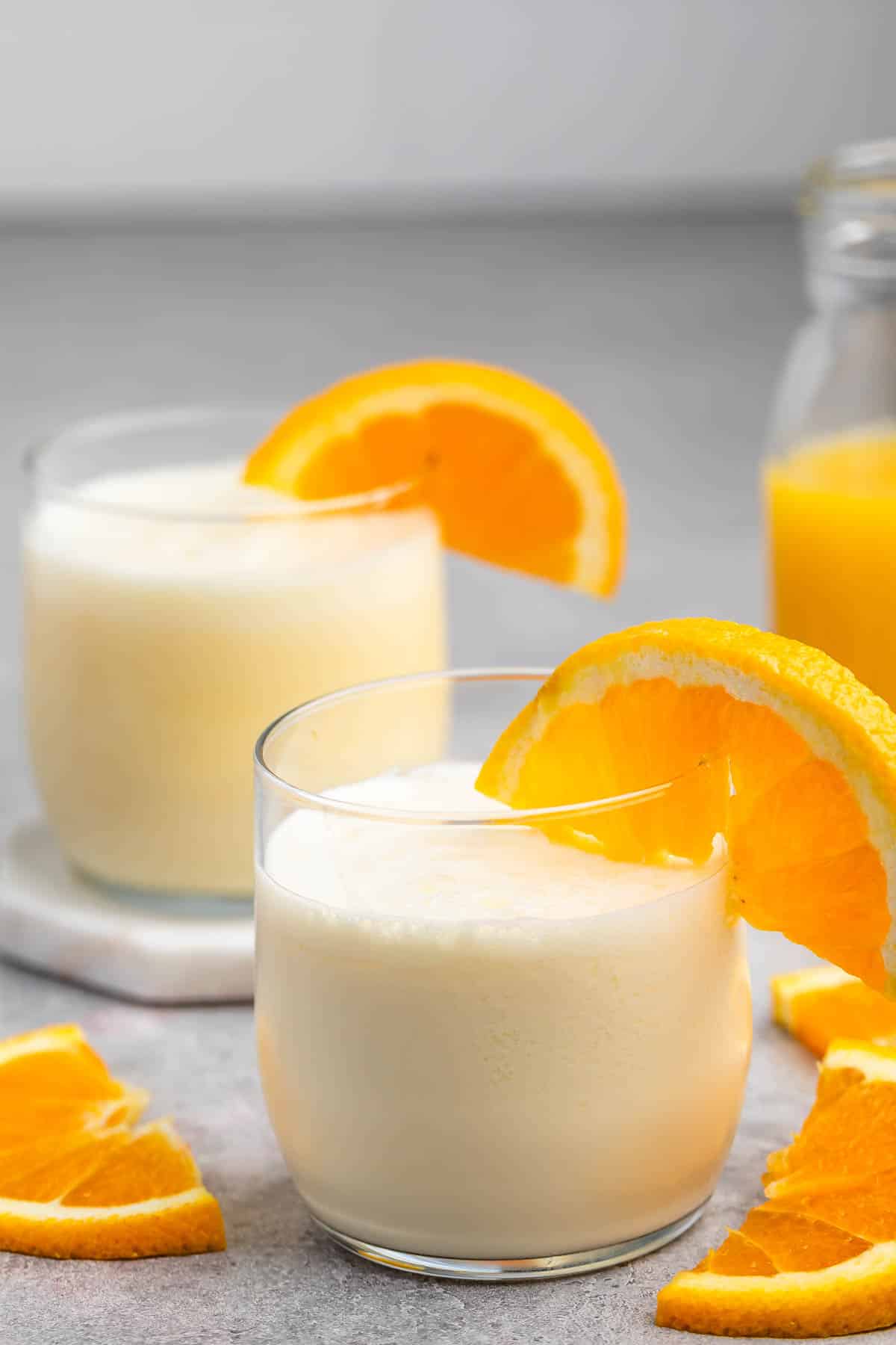 Two glasses of creamy orange smoothies garnished with orange slices. A bottle of orange juice and additional orange slices are in the background on a gray surface.