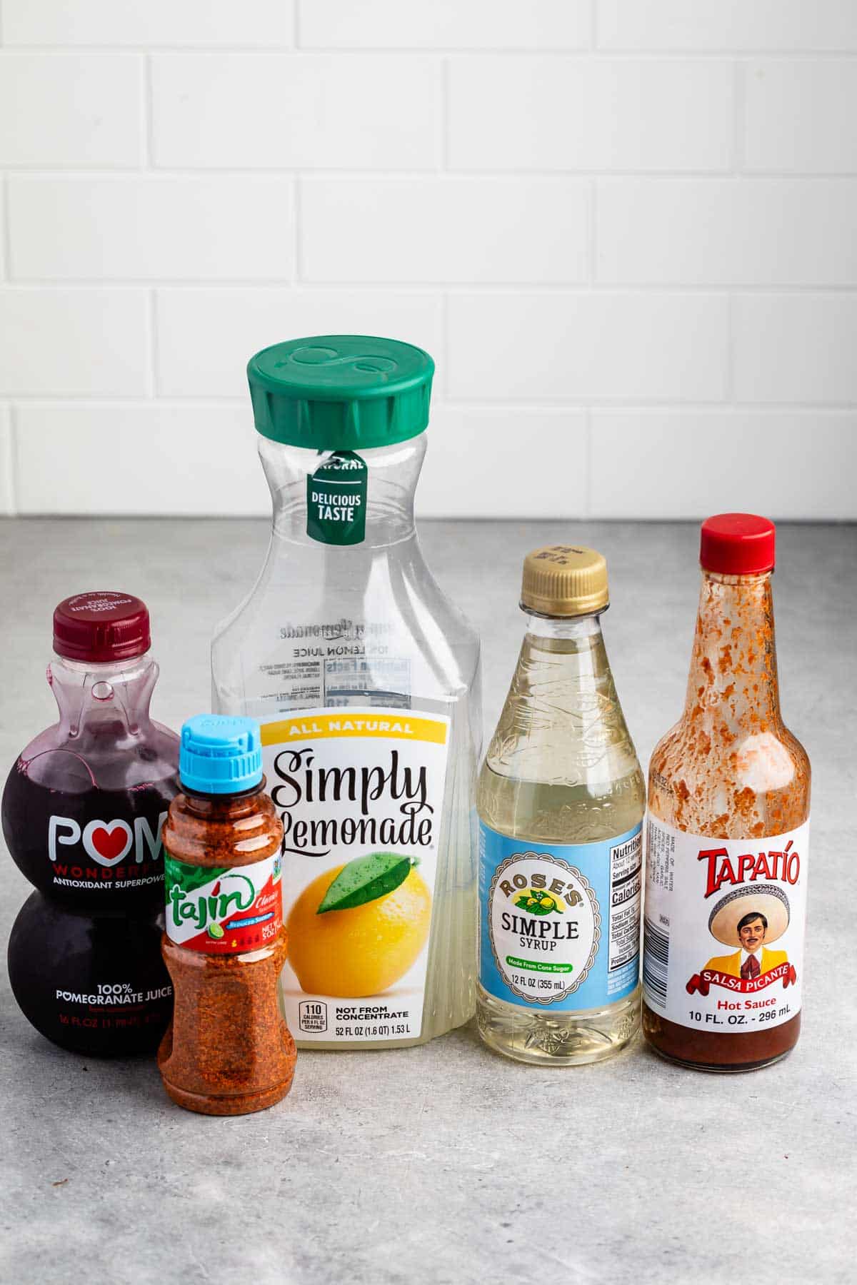 A collection of bottled products on a countertop, including pomegranate juice, lemonade, simple syrup, Tajín seasoning, and hot sauce. The background features a white tiled wall.