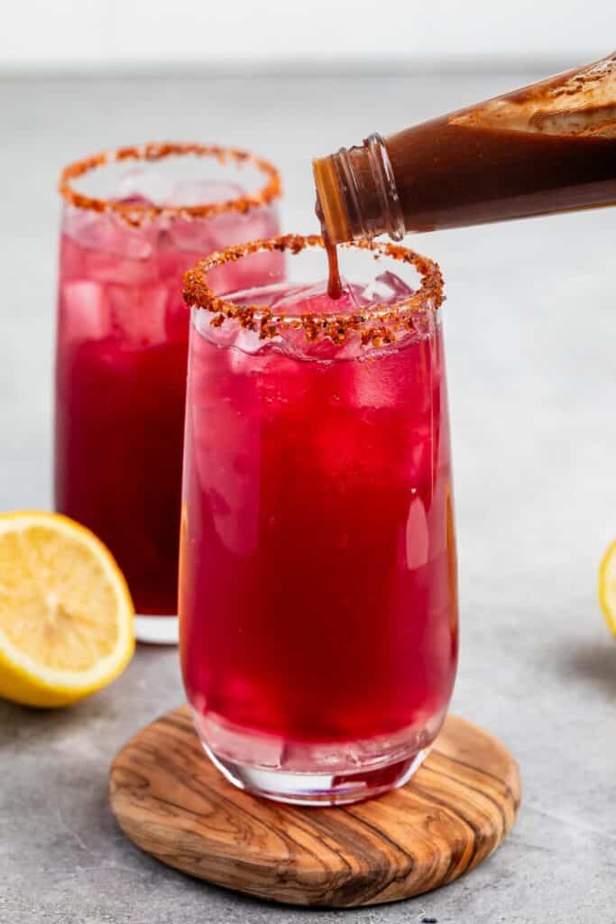 Two tall glasses filled with a bright red beverage and ice, with a chili powder rim on the glass. A bottle is pouring a brown liquid into one glass. Halved lemons are on the side, all on a wooden coaster with a grey background.