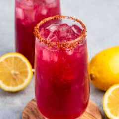 Two tall glasses filled with a bright red drink, topped with ice, on a wooden coaster. The rims are coated with a chili powder mixture. Two lemon halves and a whole lemon are placed beside the glasses on a gray surface.