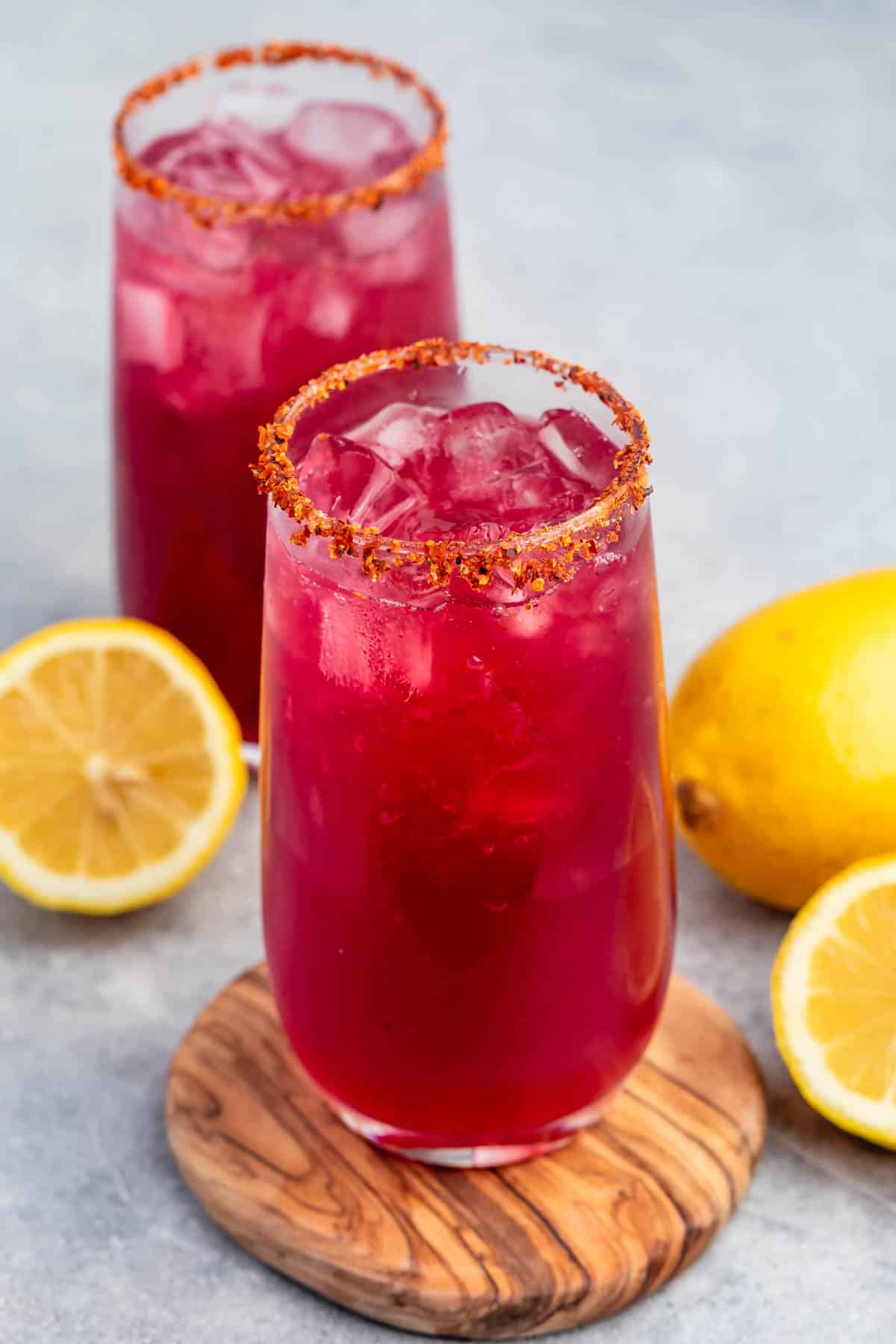 Two tall glasses filled with a bright red drink, topped with ice, on a wooden coaster. The rims are coated with a chili powder mixture. Two lemon halves and a whole lemon are placed beside the glasses on a gray surface.