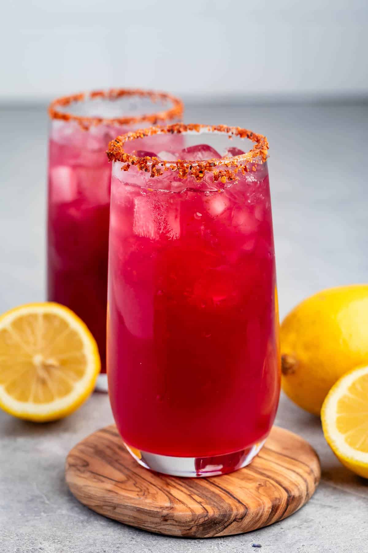 Two tall glasses of vibrant red-purple drink with ice and chili powder rims. They are placed on wooden coasters, surrounded by fresh yellow lemons, on a gray surface.