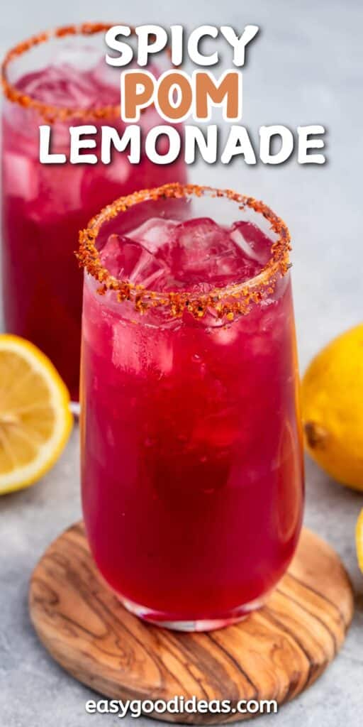 A tall glass of spicy pomegranate lemonade with a chili-rimmed edge, filled with ice and vibrant red liquid. Its placed on a wooden coaster next to a lemon half and a whole lemon. Text at the top reads Spicy Pom Lemonade.