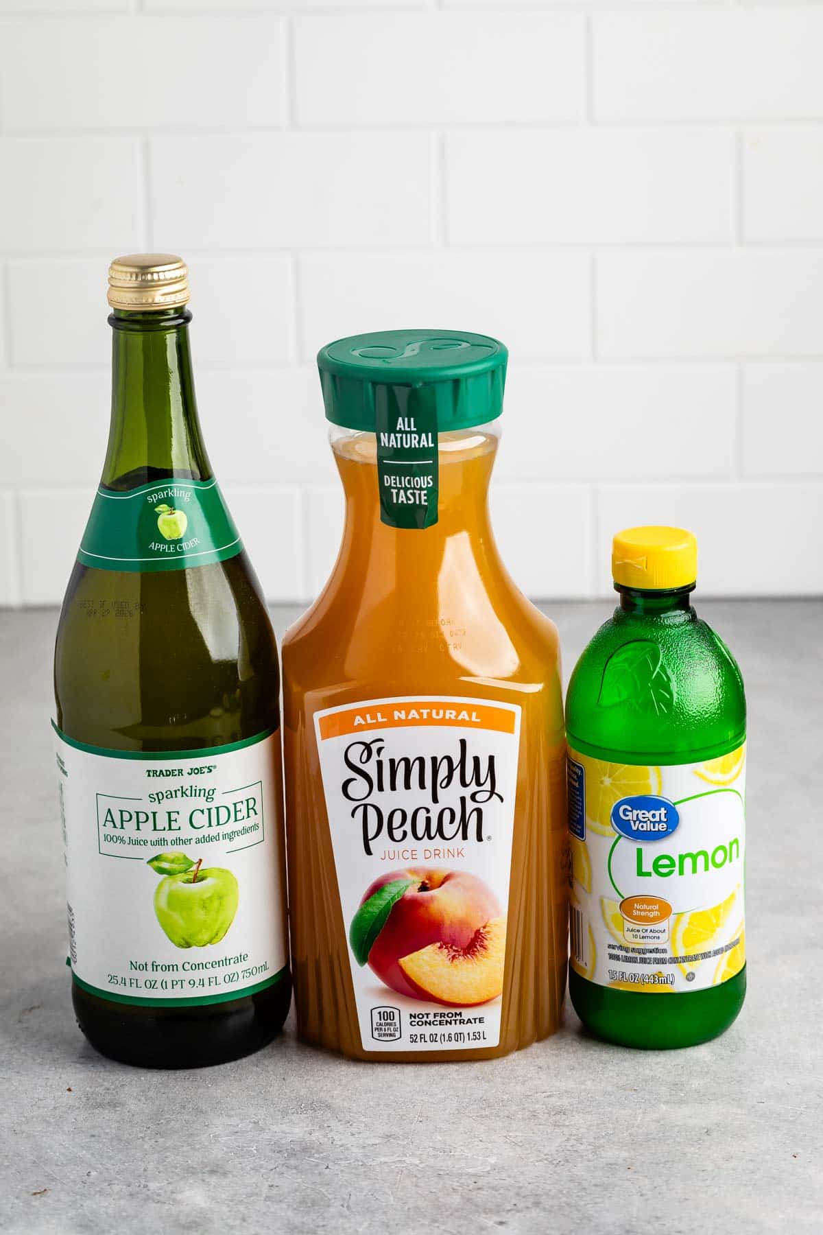 Bottles of Trader Joes sparkling apple cider, Simply Peach juice, and Great Value lemon juice are arranged side by side on a gray surface with a white tiled background.