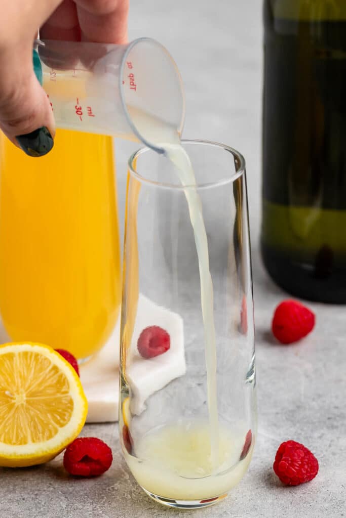 A persons hand pours a pale yellow liquid from a measuring cup into a tall glass containing raspberries. A lemon half and more raspberries are on the table. A bottle and a glass with orange liquid are in the background.