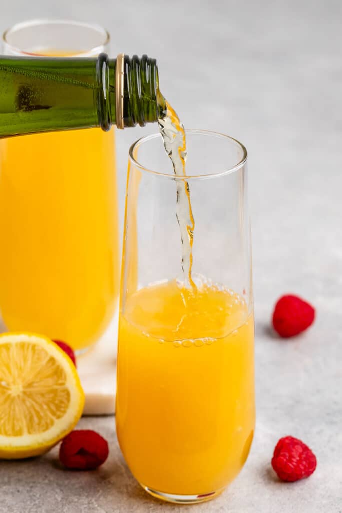 A glass of orange juice is being poured from a green bottle into a tall, clear glass. A sliced lemon and a few raspberries are scattered on the gray surface around the glass. A second filled glass is in the background.