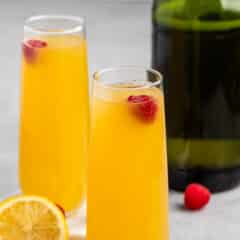 Two glasses of orange mimosa garnished with raspberries, placed on a gray surface. A sliced lemon and whole raspberries are scattered nearby, with a green bottle in the background.