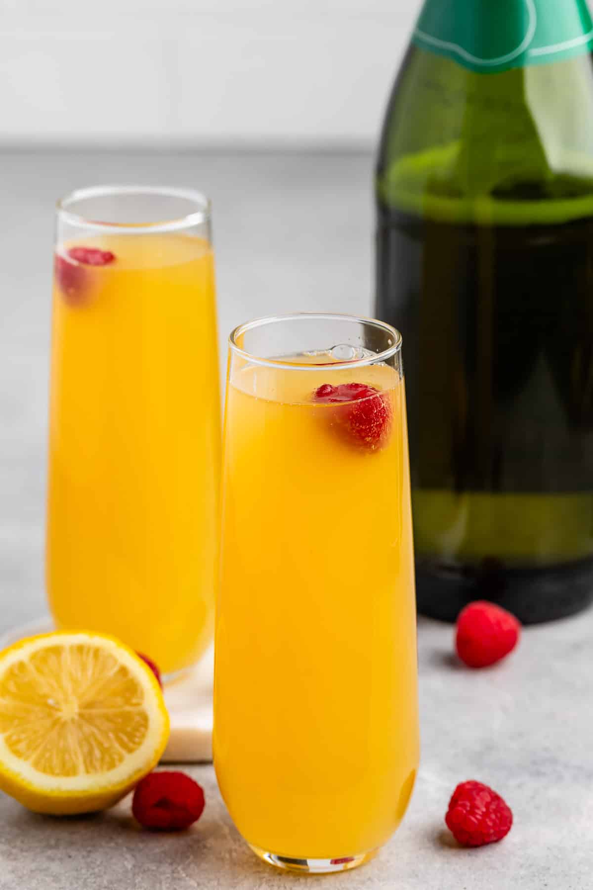 Two glasses of orange mimosa garnished with raspberries, placed on a gray surface. A sliced lemon and whole raspberries are scattered nearby, with a green bottle in the background.