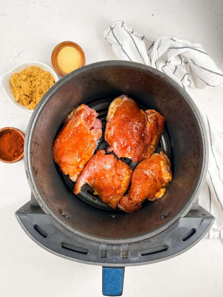 Four raw chicken thighs seasoned with a red spice rub are placed in an air fryer basket. Next to it are small bowls containing brown sugar, garlic powder, and paprika. A white dish towel is casually placed on the white countertop.