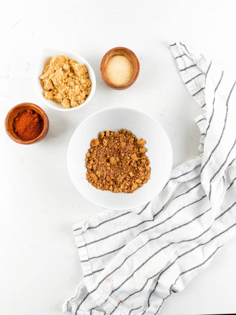 A white bowl filled with a brown crumbly mixture is surrounded by small bowls of paprika, brown sugar, and granulated sugar on a white surface. A striped kitchen towel is draped on the side.