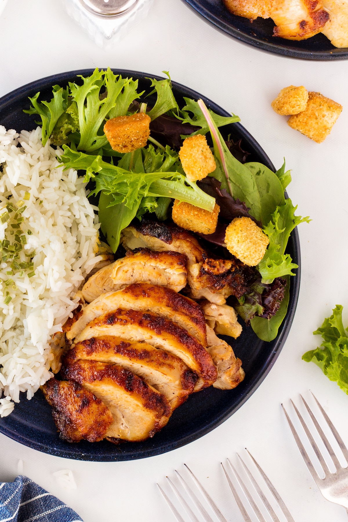 A plate with grilled chicken slices, white rice, and a green salad topped with croutons. A fork and a piece of lettuce are nearby on the light-colored table.