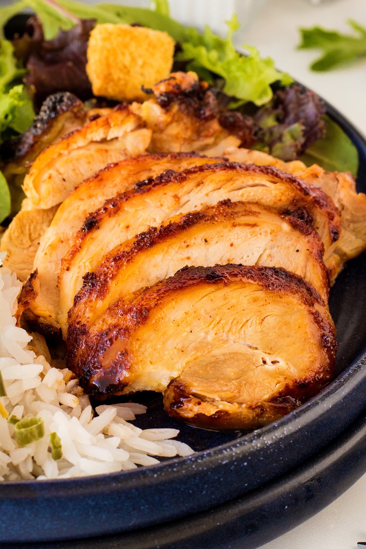 Grilled chicken slices on a plate with white rice and a fresh salad garnish, featuring lettuce and a crouton.