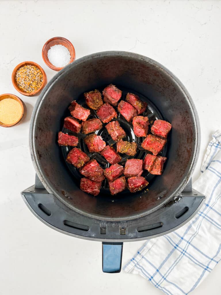 Cubes of seasoned meat in an air fryer basket, ready for cooking. Nearby are small bowls containing seasoning, salt, and breadcrumbs. A white and blue dish towel is placed on the white countertop.