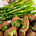 A plate of grilled steak pieces garnished with sliced green onions, served alongside a portion of grilled asparagus spears. A fork is placed on the plate beside the asparagus.